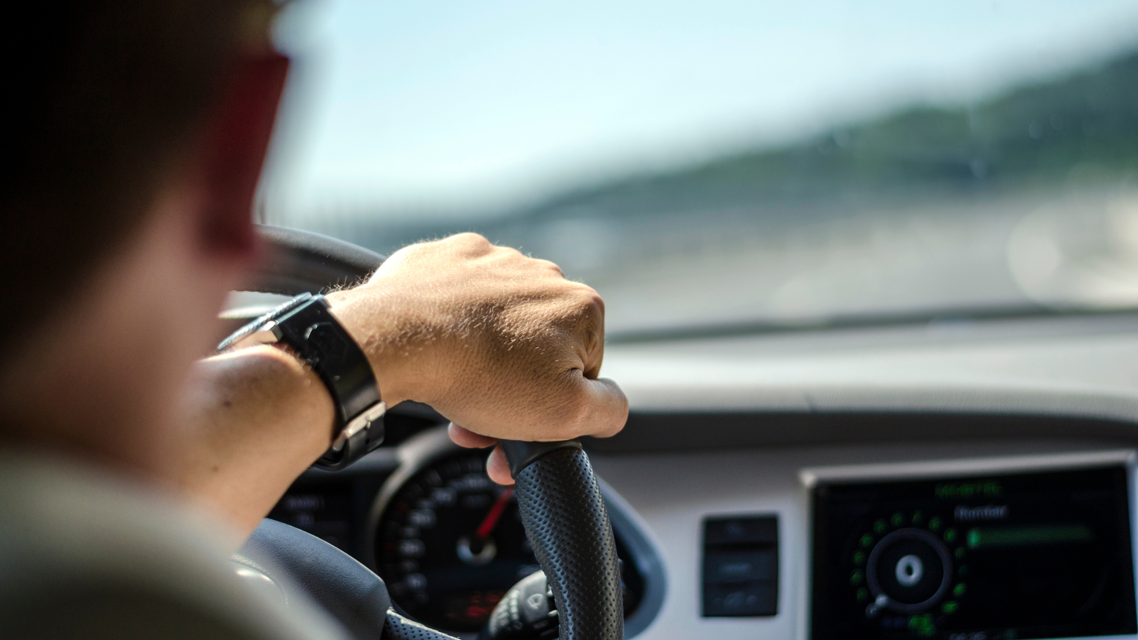 A close-up image of a person's hand gripping the steering wheel while driving a car. The dashboard and speedometer are visible in the background, slightly out of focus, along with part of the road seen through the windshield.