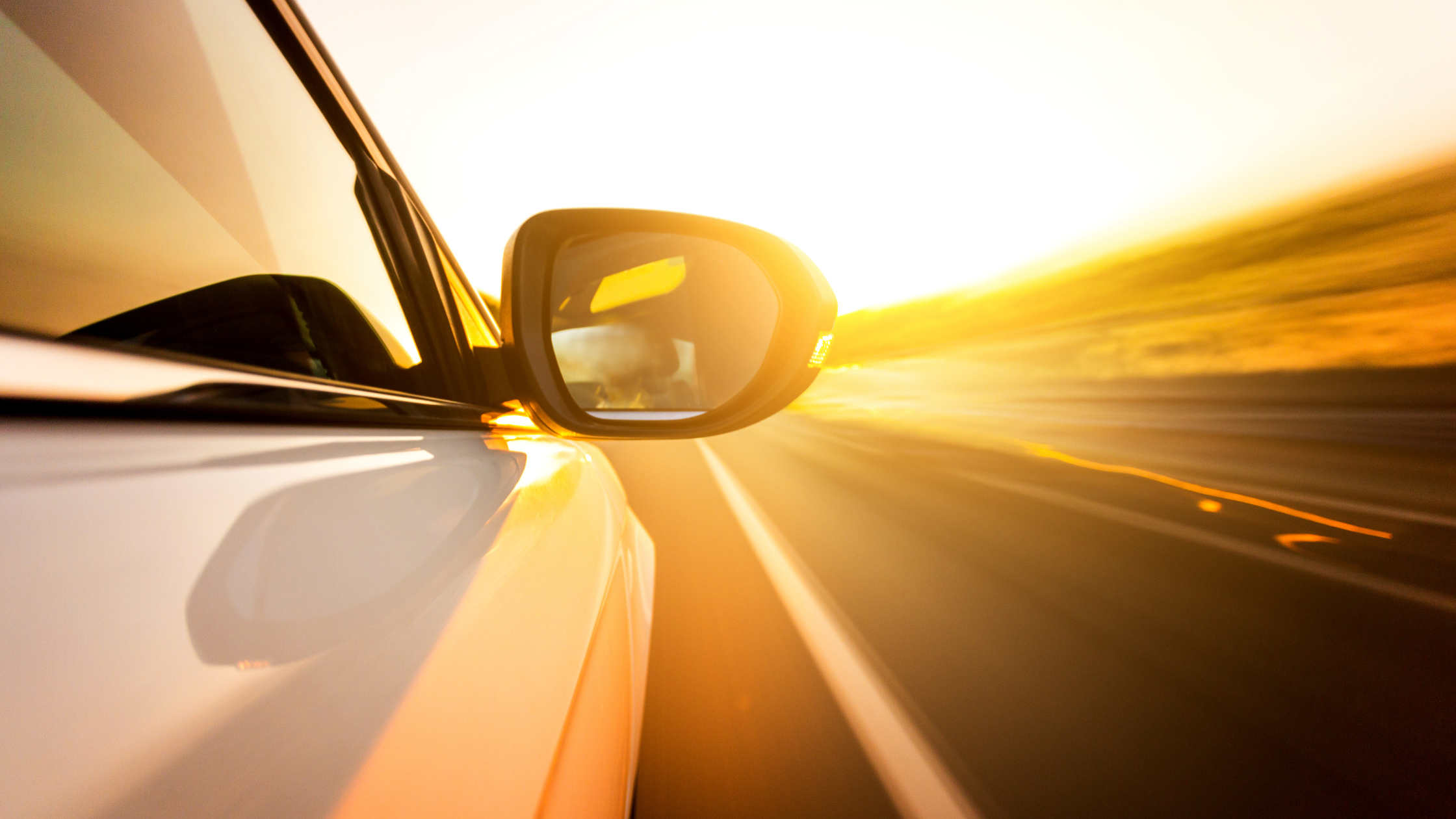 Side mirror of a white car reflecting sunlight while driving on a road at sunset.