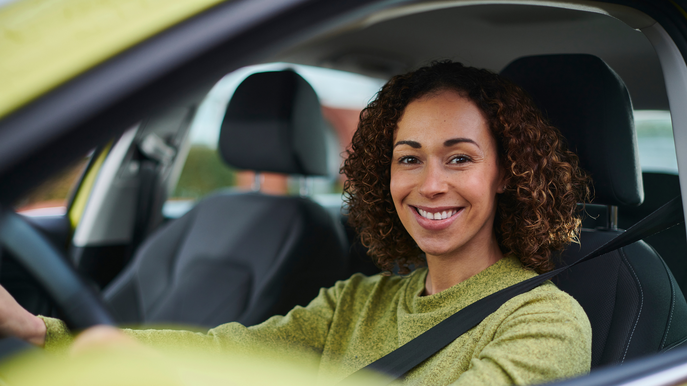 Smiling woman sitting in the driver's seat of a car, wearing a green sweater and seatbelt.